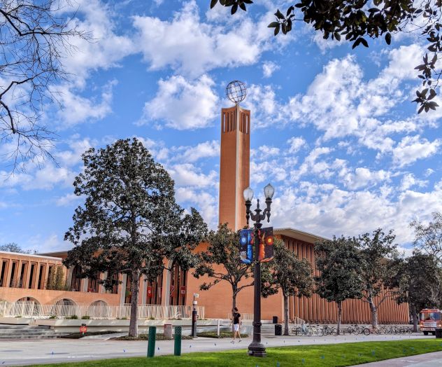 A large brick building with a tall clock tower, surrounded by trees and a clear blue sky with scattered clouds, viewed from a nearby street.