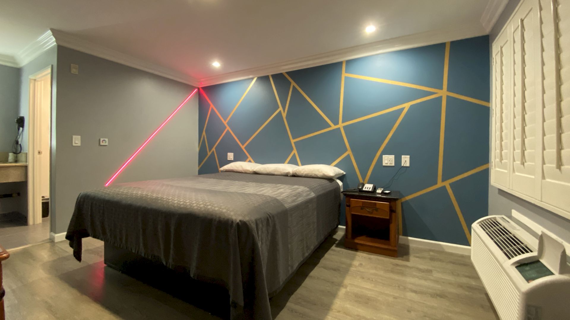 A modern bedroom with a geometric accent wall, bed with gray bedding, nightstand, air conditioner, and laser light creating a colorful effect.