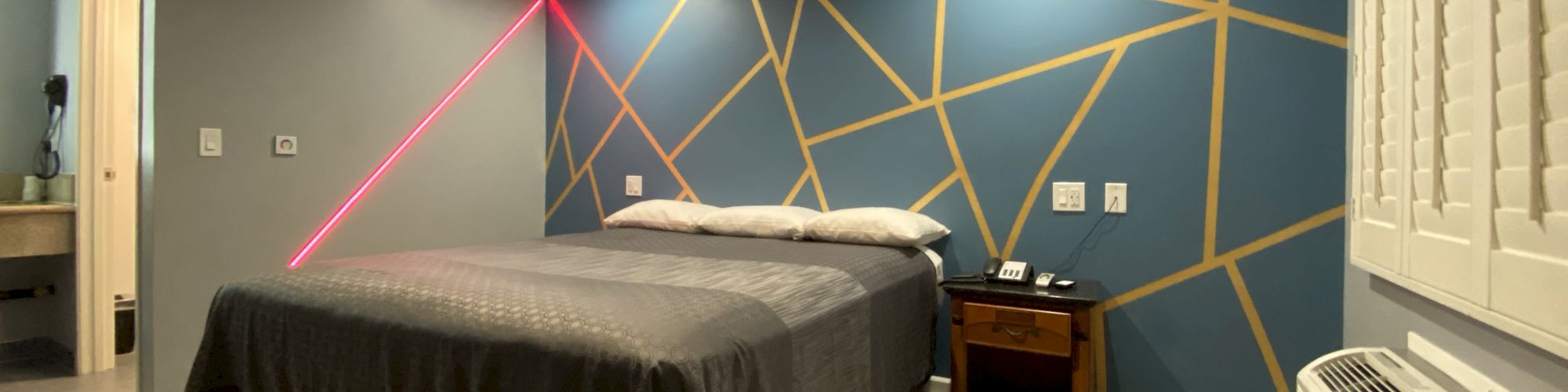 A modern bedroom with a geometric accent wall, bed with gray bedding, nightstand, air conditioner, and laser light creating a colorful effect.