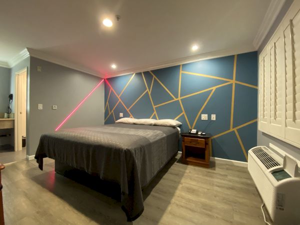 A modern bedroom with a geometric accent wall, a bed with gray bedding, two nightstands, a small desk in the corner, and an air conditioning unit.