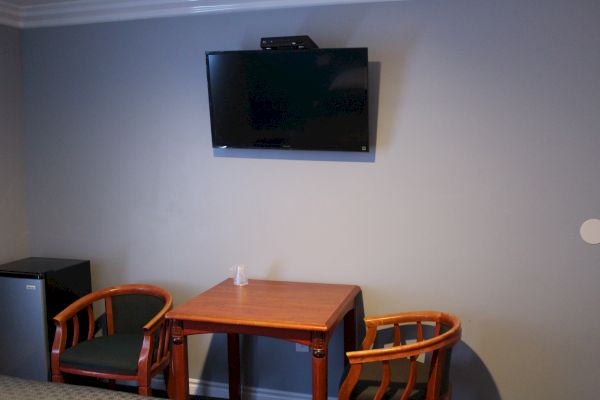 A room with a wall-mounted TV, a wooden table with two chairs, and a mini-fridge in the corner under grey walls.