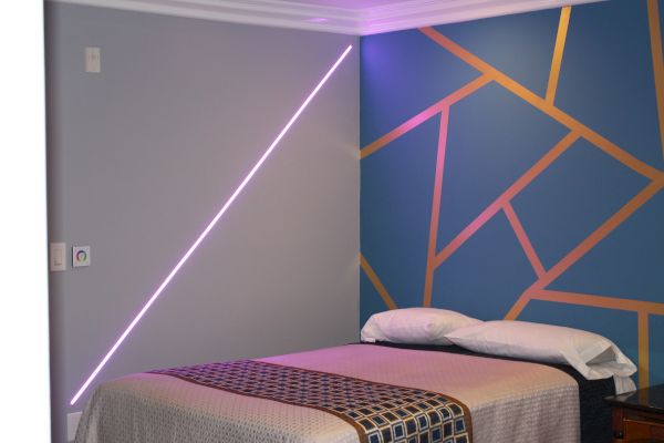 A neatly made bed with a patterned bedspread, colorful geometric accent wall design, and a diagonal pink neon light on a plain gray wall.