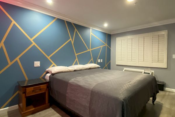 A modern bedroom with a geometric accent wall, a bed with gray bedding, nightstands, recessed lighting, and wooden flooring.