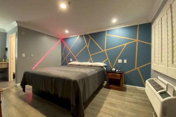 This image shows a modern bedroom with a geometric accent wall, a bed with a gray bedspread, a nightstand, a desk in the corner, and a window unit air conditioner.