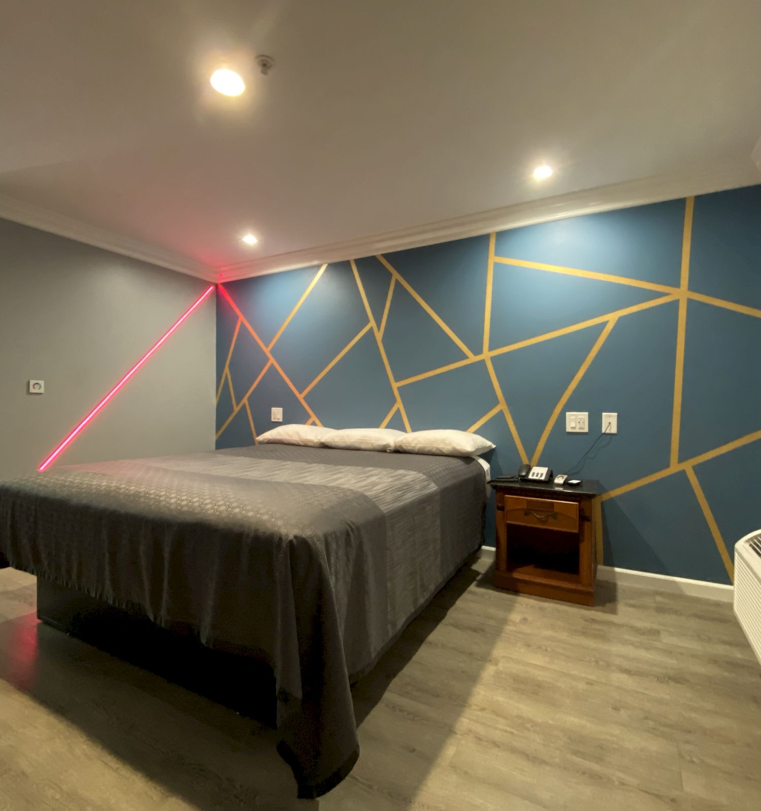 A modern bedroom with a geometric accent wall, bed, nightstand, air conditioner, and desk space in the corner.