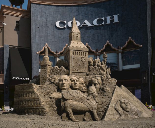 A detailed sand sculpture features landmarks and faces, including a pyramid, possibly Mount Rushmore, and the Colosseum, in front of Coach store.