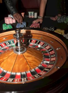 The image shows a roulette table with a roulette wheel in the foreground, and people handling poker chips in the background.