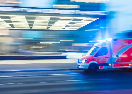 The image shows a red and white ambulance with flashing lights moving quickly on a street, indicating an emergency situation.