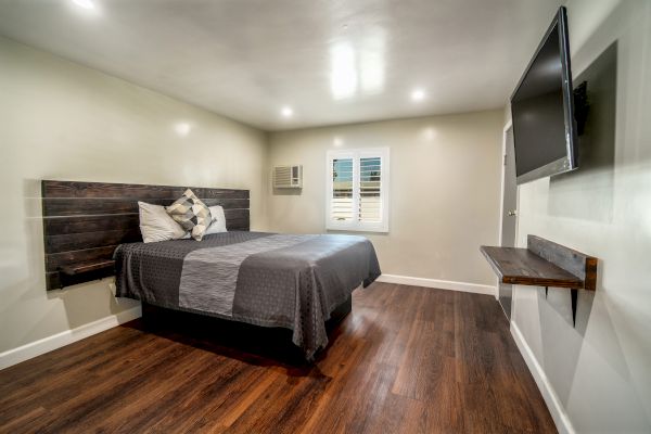 A modern bedroom with wooden flooring, a bed with gray linens, a wall-mounted TV, a small shelf, and a window with shutters.