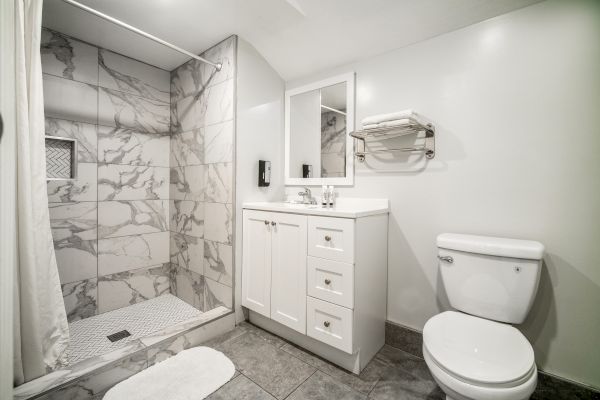 A modern bathroom with a marble-tiled shower, a white vanity with drawers, a toilet, and a wall-mounted towel rack under a mirror.