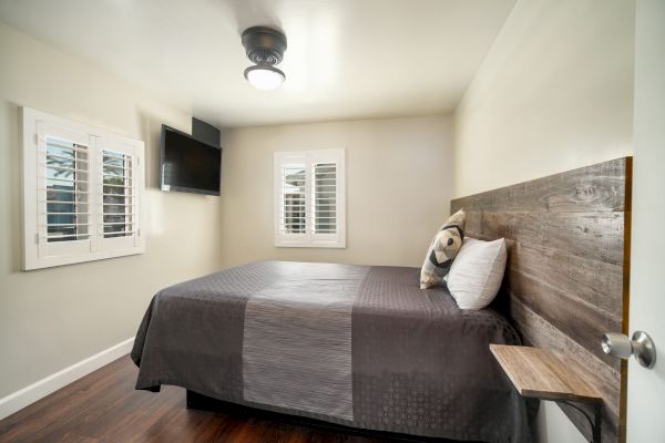 A small bedroom with a bed, wooden headboard, pillow, wall-mounted TV, and two windows with white shutters, on a wooden floor with a ceiling light.