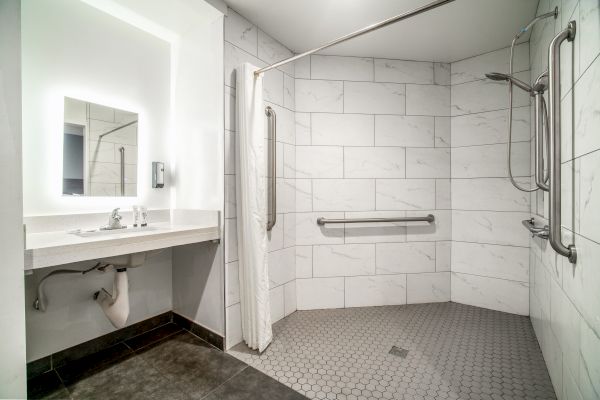 An accessible bathroom with a roll-in shower, white tiles, grab bars, a sink with a mirror, and a shower curtain.