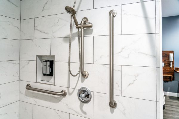 A shower with a handheld showerhead, grab bars, and a built-in soap dispenser against a white tiled wall.