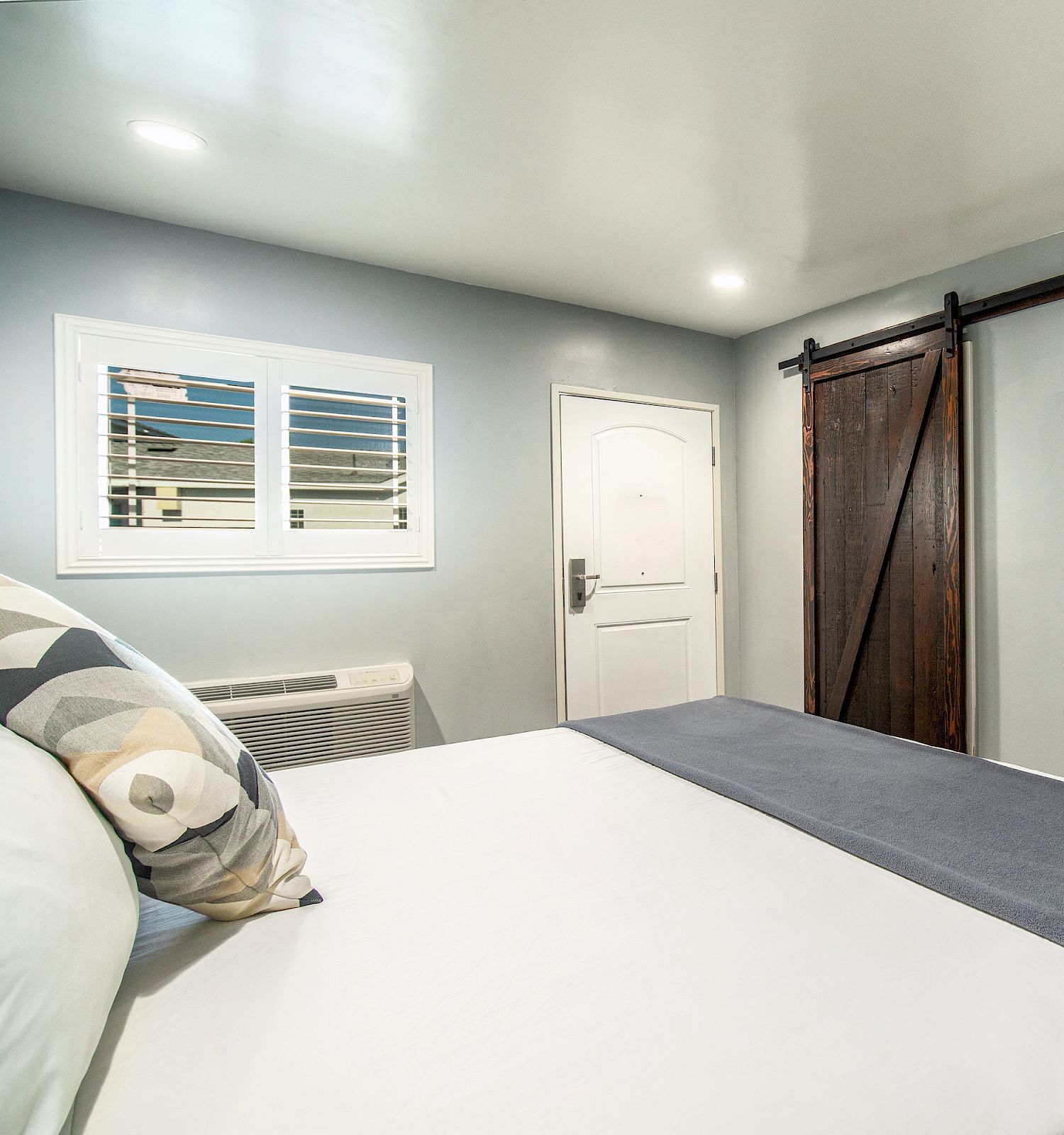 A modern bedroom with a large bed, wall-mounted TV, barn-style door, air conditioning unit, and white door under a window with shutters.