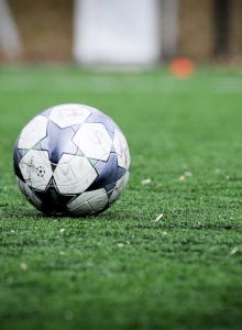 A soccer ball rests on a green field, with blurred goalposts and background elements visible, indicating an outdoor sports environment.