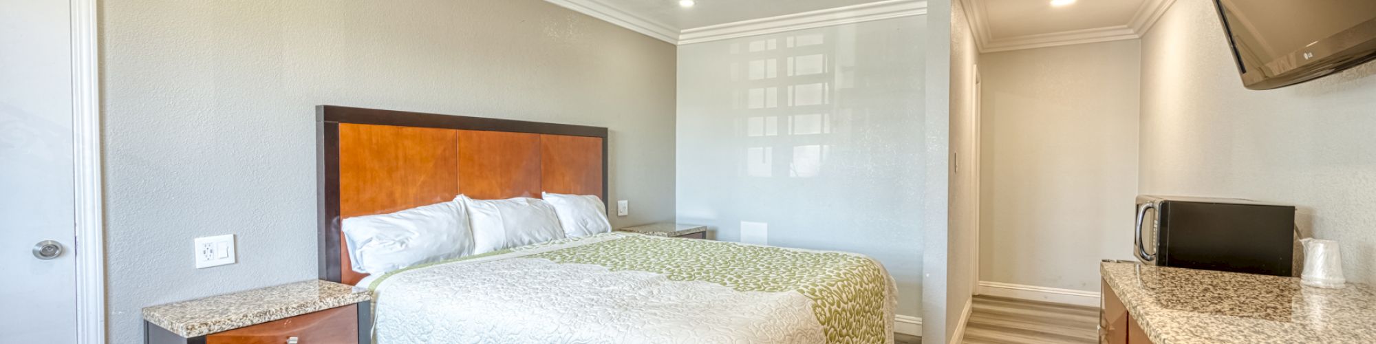 A modern hotel room with a king-sized bed, nightstand, mounted TV, microwave, and a dresser. The floor is wood, and the walls are light colored.