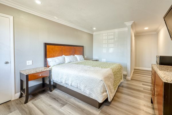 The image shows a bright hotel room with a queen bed, a side table, a chest of drawers, a wall-mounted TV, and a microwave.