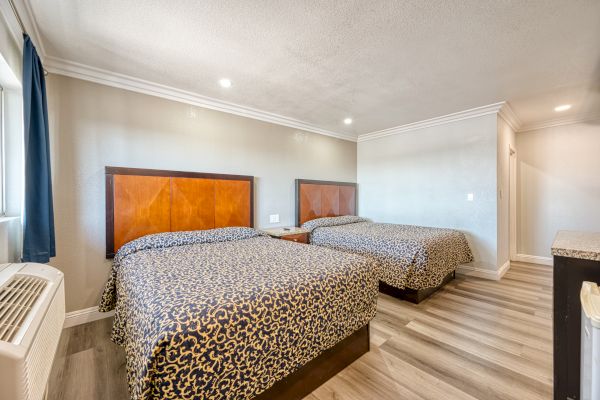 A hotel room with two double beds, patterned bedspreads, a nightstand, air conditioner, wood flooring, and white walls.