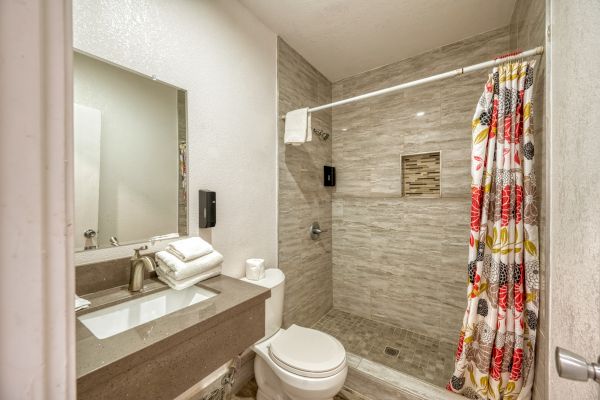 The image shows a modern bathroom with a sink, mirror, toilet, and a walk-in shower with colorful curtain and grey tiling.