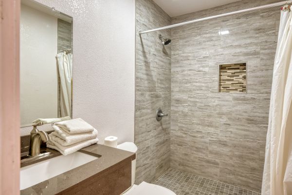 The image shows a modern bathroom with a walk-in shower featuring grey tiles and a recessed shelf, a countertop with folded towels, and a toilet paper roll.