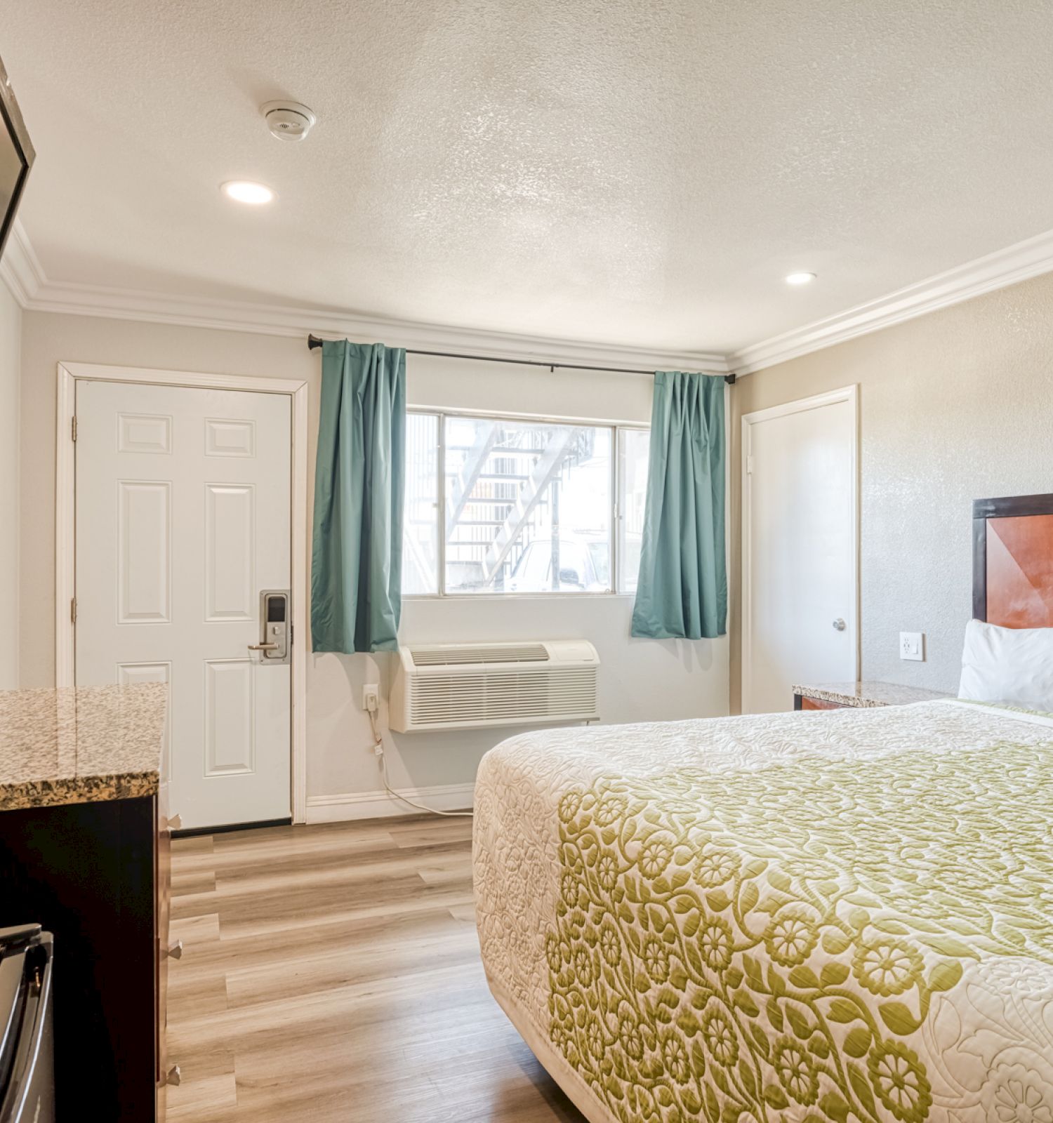 The image shows a clean, simple hotel room with a bed, microwave, mounted TV, window with curtains, an air conditioning unit, and a door.