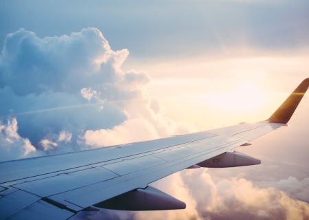 This image shows the wing of an airplane flying above the clouds, with the sun shining in the background, creating a scenic view.