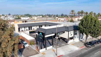 An aerial view of a small motel with a parking area, situated on a street lined with trees and surrounded by other buildings.