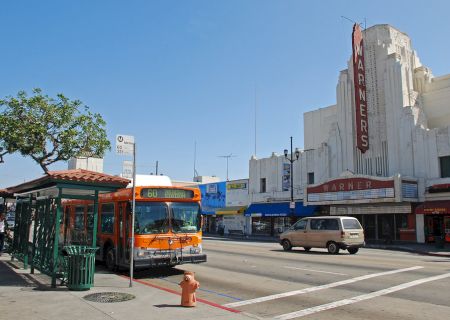 A street scene features a bus at a stop, a theater with a vertical marquee, storefronts, and a tree, under a clear blue sky.