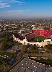 Aerial view of a large stadium with red seats, surrounded by an urban landscape and greenery, under a clear blue sky.