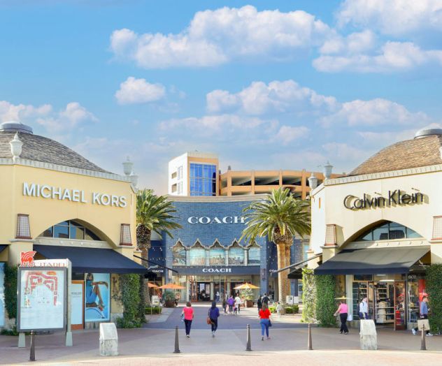 An outdoor shopping center with Michael Kors, Coach, and Calvin Klein stores under a partly cloudy sky. People are walking around.