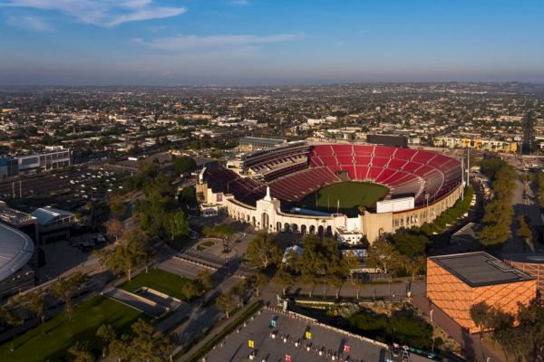 The image shows an aerial view of a large stadium with red seats, surrounded by a cityscape under a blue sky.