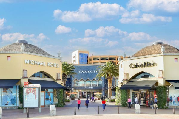 The image shows an outdoor shopping area with stores under blue skies, including Michael Kors, Coach, and Calvin Klein, with palm trees nearby.