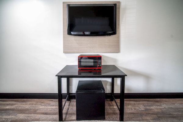 The image shows a mounted TV, a red microwave on a table, and a small fridge underneath, against a plain wall.