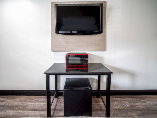 The image shows a mounted TV, a red microwave on a table, and a small fridge underneath, against a plain wall.