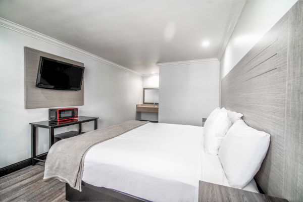 A modern hotel room with a neatly made bed, wall-mounted TV, table with items, and a mirror at the far end. White and gray tones dominate.