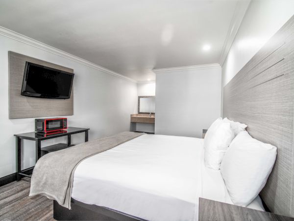 A modern hotel room with a neatly made bed, wall-mounted TV, table with items, and a mirror at the far end. White and gray tones dominate.