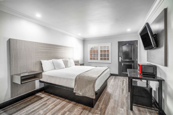 A modern hotel room with a bed, wall-mounted TV, and desk. The room has wooden flooring, white walls, and a window with blinds.