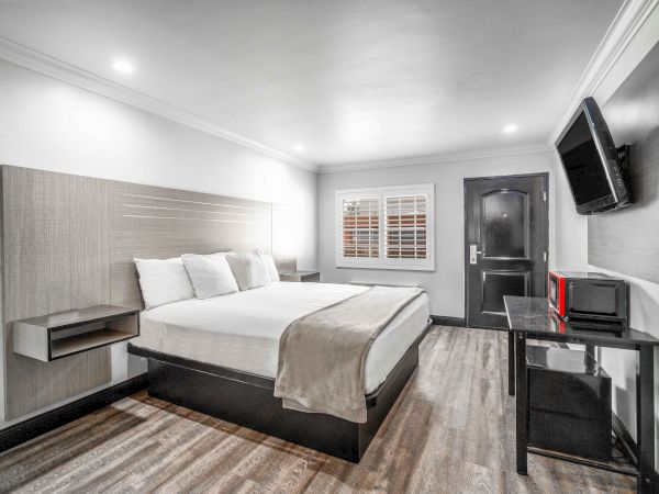 A modern hotel room with a bed, wall-mounted TV, and desk. The room has wooden flooring, white walls, and a window with blinds.