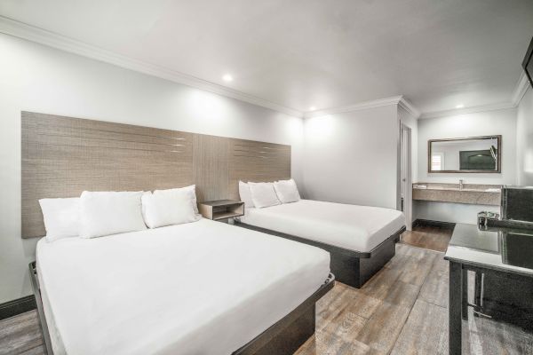 The image shows a hotel room with two beds, a vanity area with a mirror, and a TV on a stand, featuring modern and minimalist decor.