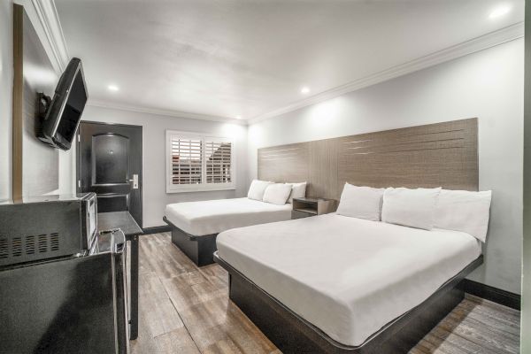 The image shows a modern hotel room with two beds, a wall-mounted TV, microwave, and a window with shutters, featuring clean decor.