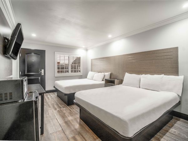 The image shows a modern hotel room with two beds, a wall-mounted TV, microwave, and a window with shutters, featuring clean decor.