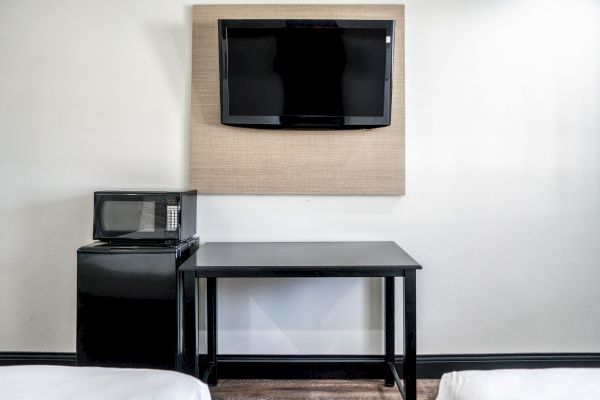 The image shows a wall-mounted TV, a small black table, a microwave, and a mini fridge in a room with white walls.