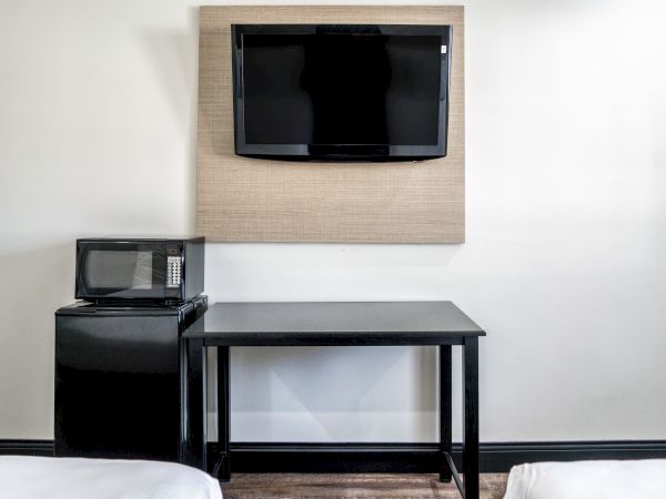 The image shows a wall-mounted TV, a small black table, a microwave, and a mini fridge in a room with white walls.
