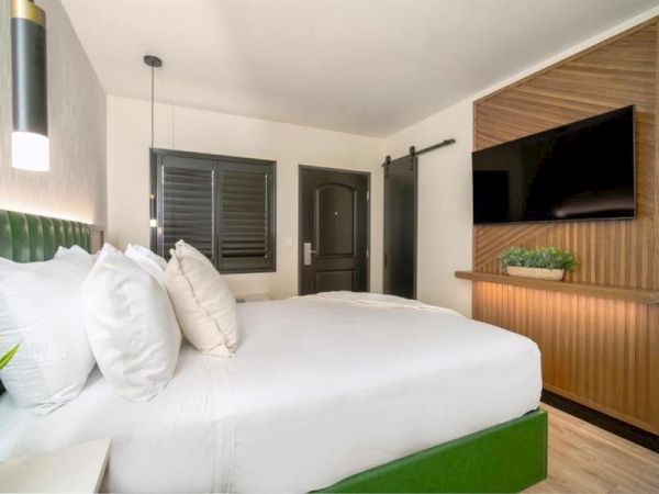 The image shows a modern hotel room with a neatly made bed, pillows, a TV mounted on the wall, and a sliding door.