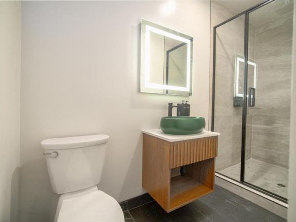 A modern bathroom features a toilet, a vanity with a green sink and mirror, and a glass-enclosed shower on gray tiled flooring.