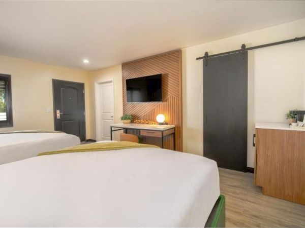 The image shows a modern hotel room with two beds, a TV on the wall, a sliding door, and a counter with a coffee maker.
