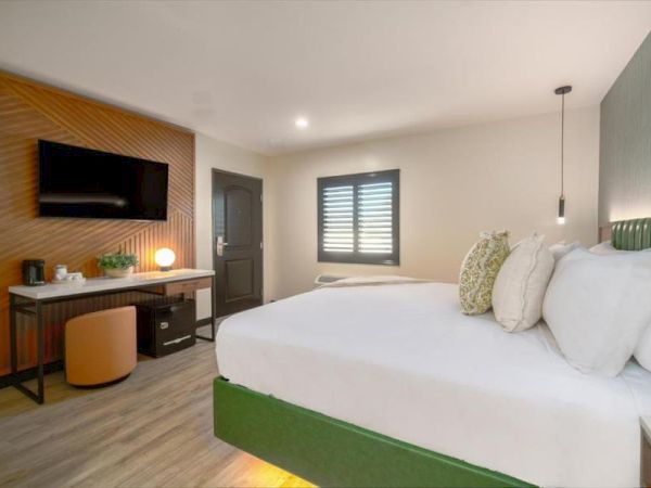 A modern hotel room features a large bed, wall-mounted TV, desk with lamp, chair, and window with shutters. The design is sleek and minimalistic.