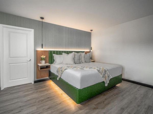 A modern bedroom with a large bed, green accents, wood flooring, stylish lighting, and a side table by a closed white door completes the scene.