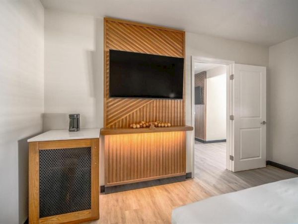 The image shows a modern hotel room with a wall-mounted TV, wooden paneling, a small cabinet, and an open doorway leading to another room.