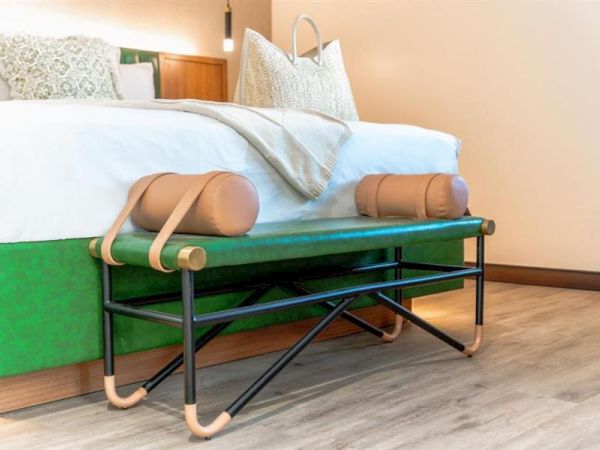 The image shows a bedroom with a neatly made bed, decorative pillows, and a stylish bench with cylinder cushions at the foot of the bed.